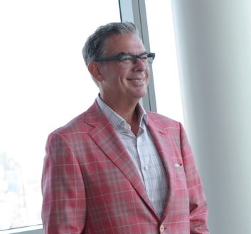 Elvis Duran standing in a city office in jacket and open-neck shirt
