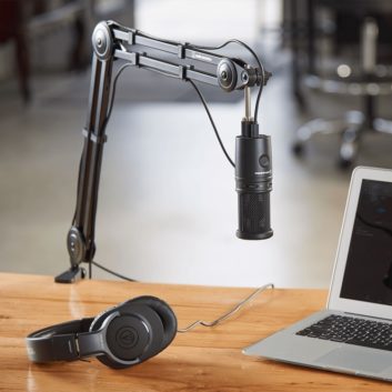 An Audio-Technica AT8700 microphone boom arm shown holding a microphone at a content creator's work area, also equipped with laptop and headphones