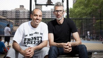 Adrian “Stretch Armstrong” Bartos and Bobbito García sit on a bench next to a basketball court