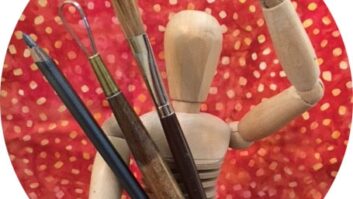 A wooden modeling figure holds paint brushes and appears to wave at the camera.