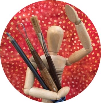 A wooden modeling figure holds paint brushes and appears to wave at the camera.