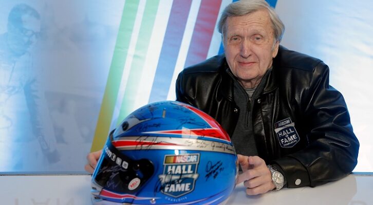 Ken Squire sitting at a table with a racing helmet in front of him on the table bearing the signatures of NASCAR drivers.