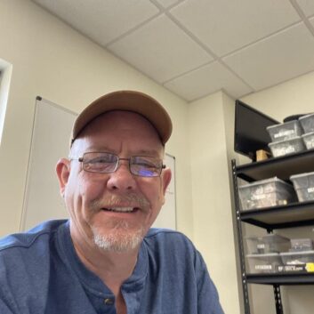 A man in a blue work shirt and baseball cap smiles at the camera. His sitting in a room with shelves of equipment and supplies behind him.