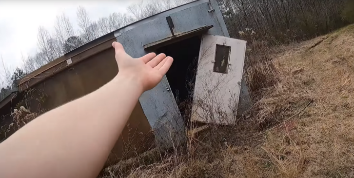 A still from a YouTube video showing a dilapidated equipment shelter in an overgrown field, with the camera operator's hand gesturing in front of the lens