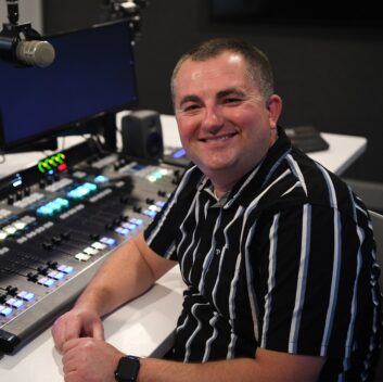 A man sits in a radio studio with a console and microphone behind him, smiling at the camera, and wearing a short-sleeved striped shirt