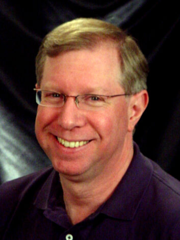 A man wearing glasses and a casual shirt smiles at the camera.