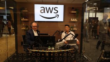Two people sit in a radio studio enclosed by glass on the convention floor of the NAB Show, under a sign with the AWS logo