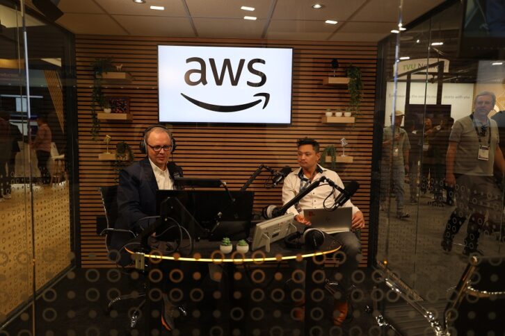 Two people sit in a radio studio enclosed by glass on the convention floor of the NAB Show, under a sign with the AWS logo