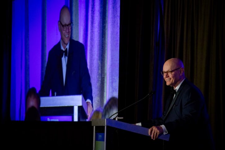 A man speaks at a podium in a darkened auditorium. An image of him is visible on a large screen behind him.