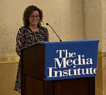 A woman stands at a lecturn, smiling and looking down at her notes. The lecturn has a sign that reads "The Media Institute"