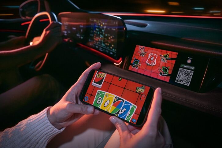 A view of a car dashboard, with a passenger's hands in the foreground holding a smartphone. The phone and dashboard both display a video game