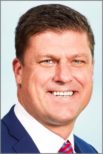 A headshot of a man smiling, wearing a blue suit jacket and red tie