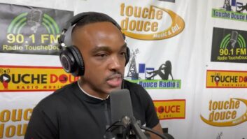 A man talks into a microphone while wearing headphones. Logos of Radio Touche Douce are on the wall behind him.