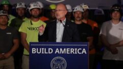 FCC Chairman Brendan Carr stands at a podium in a still from a YouTube video. Tower workers in T shirts and safety helmets stand behind him.