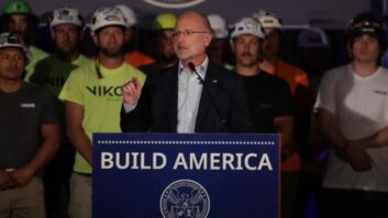 FCC Chairman Brendan Carr stands at a podium in a still from a YouTube video. Tower workers in T shirts and safety helmets stand behind him.