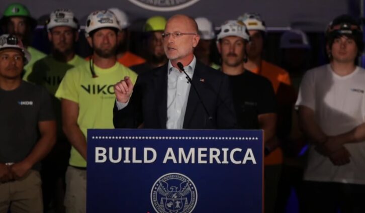 FCC Chairman Brendan Carr stands at a podium in a still from a YouTube video. Tower workers in T shirts and safety helmets stand behind him.