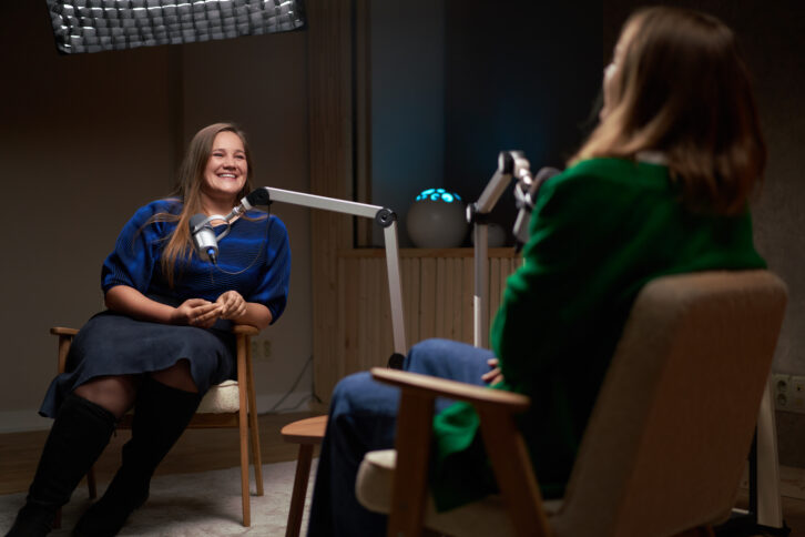Smiling female podcast host sitting on chair and listening to funny story from guest in modern studio 