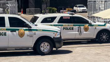 Two Miami-Dade police K-9 cars are parked near a metal security fence on a bright sunny day.