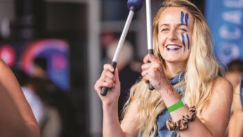 A woman wearing face paint holds two drumming sticks in the air