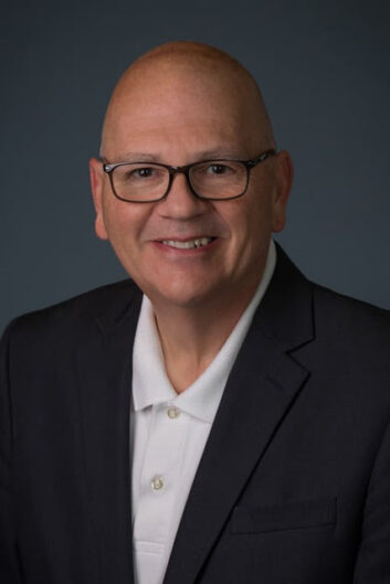 A man wearing a business jacket and polo shirt smiles at the viewer.