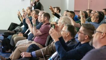 A group of people seated in a conference room aim their phones and cameras at the podium, which is out of our frame of vision to the left