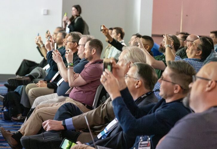 A group of people seated in a conference room aim their phones and cameras at the podium, which is out of our frame of vision to the left