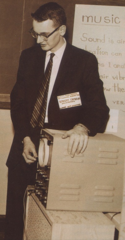 A black and white photo of a young man in sut and tie with an oscilloscope, standing in front of a lecture board