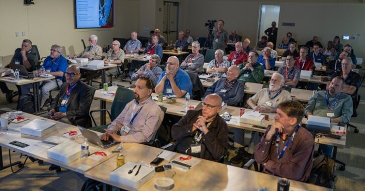 A room full of people sitting at conference tables listening to a speaker who is not visible to us.