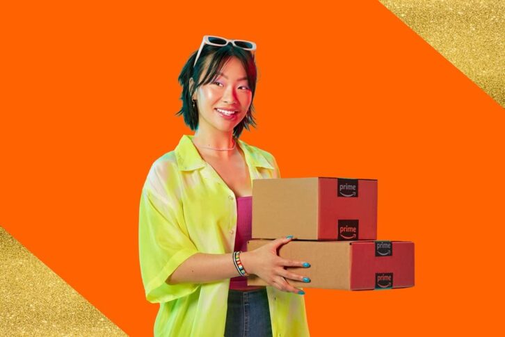 A woman in casual clothing smiles while holidng two boxes with the Amazon logo.