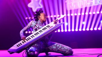 A person dressed in a colorful spandex outfit kneels on a stage playing a keyboard like a guitar, bathed in purple light