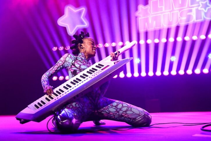 A person dressed in a colorful spandex outfit kneels on a stage playing a keyboard like a guitar, bathed in purple light