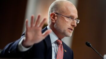 A man in a suit and tie sits at a hearing table and gestures with his right hand
