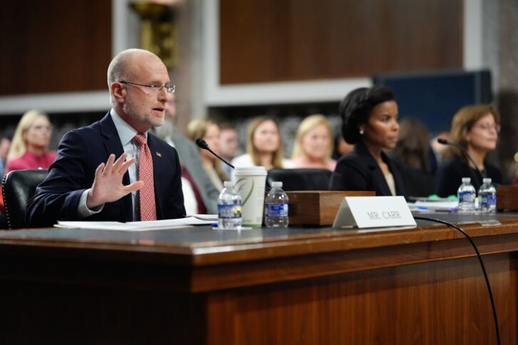 A man and two women in business clothes sit at a hearing table in Washington. The man is speaking into a microphone.
