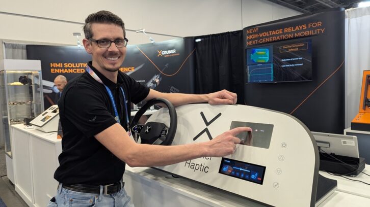 A man in the Gruner booth touches a mock dashboard display while smiling at the camera