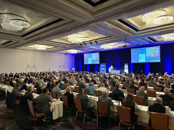 A large conference room is filled with seated attendees listening to a presentation