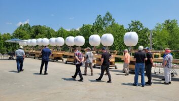 A 13-bay FM antenna with radomes lies on its side on the ground at a Dielectric factory event, as visitors walk past to inspect it