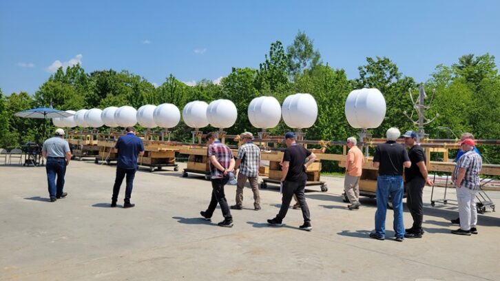 A 13-bay FM antenna with radomes lies on its side on the ground at a Dielectric factory event, as visitors walk past to inspect it