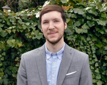 Kyle Suess stands outside in front of shrubbery, wearing a sports jacket and open-collared shirt