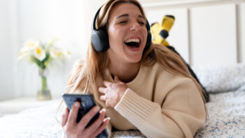 Young adult woman laughing enjoying content with headphones and smartphone in bedroom