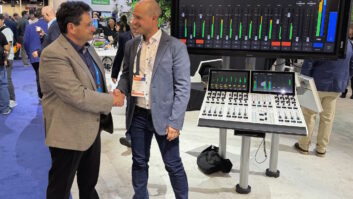Two men shake hands in a trade show booth with a mixing console nearby