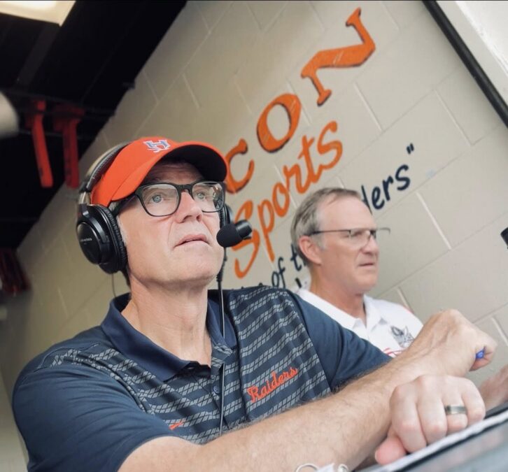Two men sit in a broadcast booth looking toward a sports field