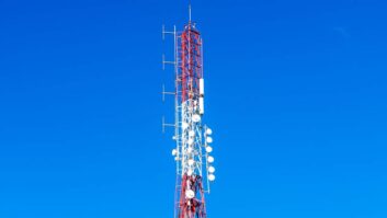 Top section of a red and white communication tower with antennas, dish units, and metal lattice, rising into a clear deep blue sky.