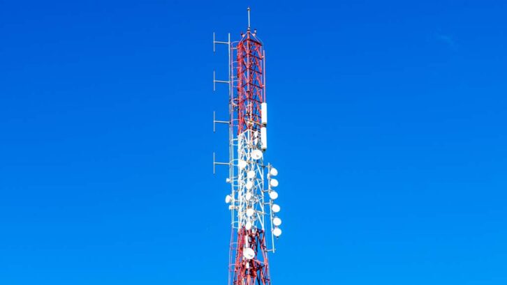 Top section of a red and white communication tower with antennas, dish units, and metal lattice, rising into a clear deep blue sky.