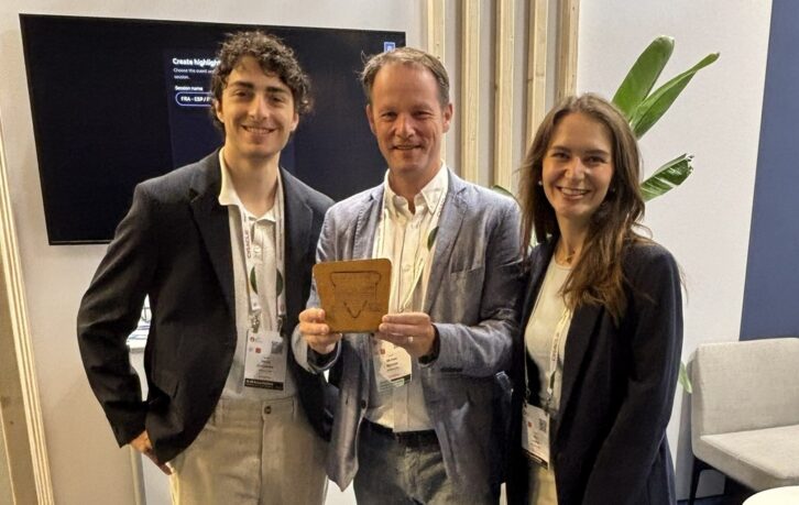 Three people stand in the deliver.media booth at IBC, one holding an award