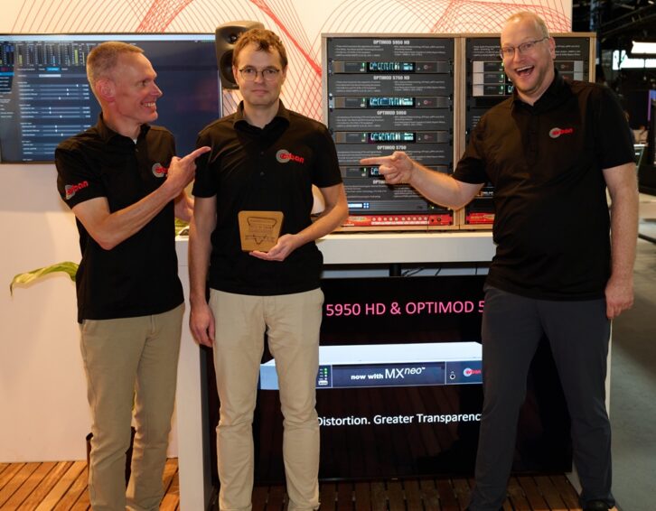 Three people stand in the Orban booth at IBC with an award. Two are pointing at the third in a humorous pose