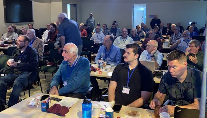 People sit and stand in a conference room listening to a presenter not visible in the photo