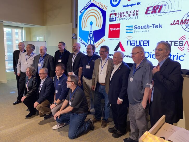 14 people stand in front of a large projection of sponsor logos in a conference room