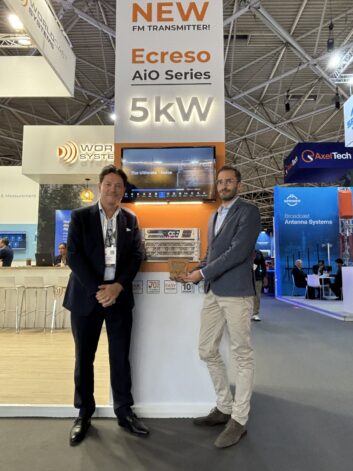 Two men stand in the WorldCast Systems booth at IBC, one holds an award